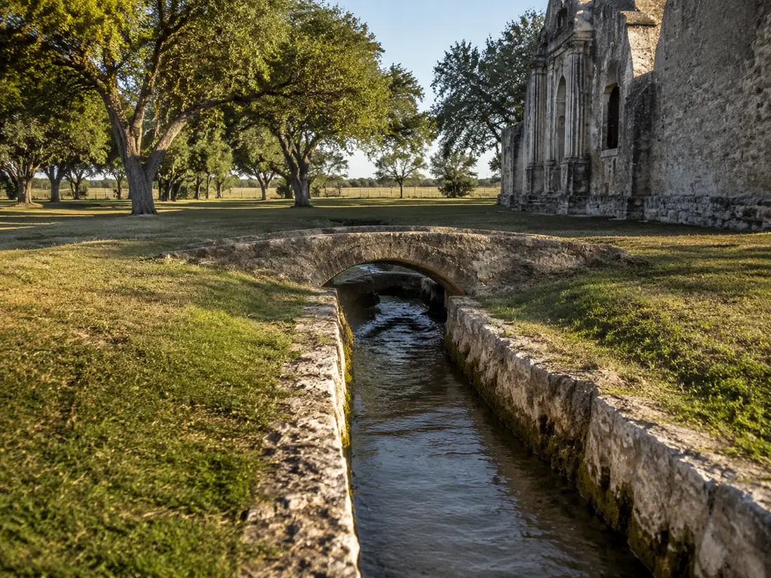 A stone-lined acequia running low and clear between grass banks &mdash; the colonial irrigation channel that made farming here possible